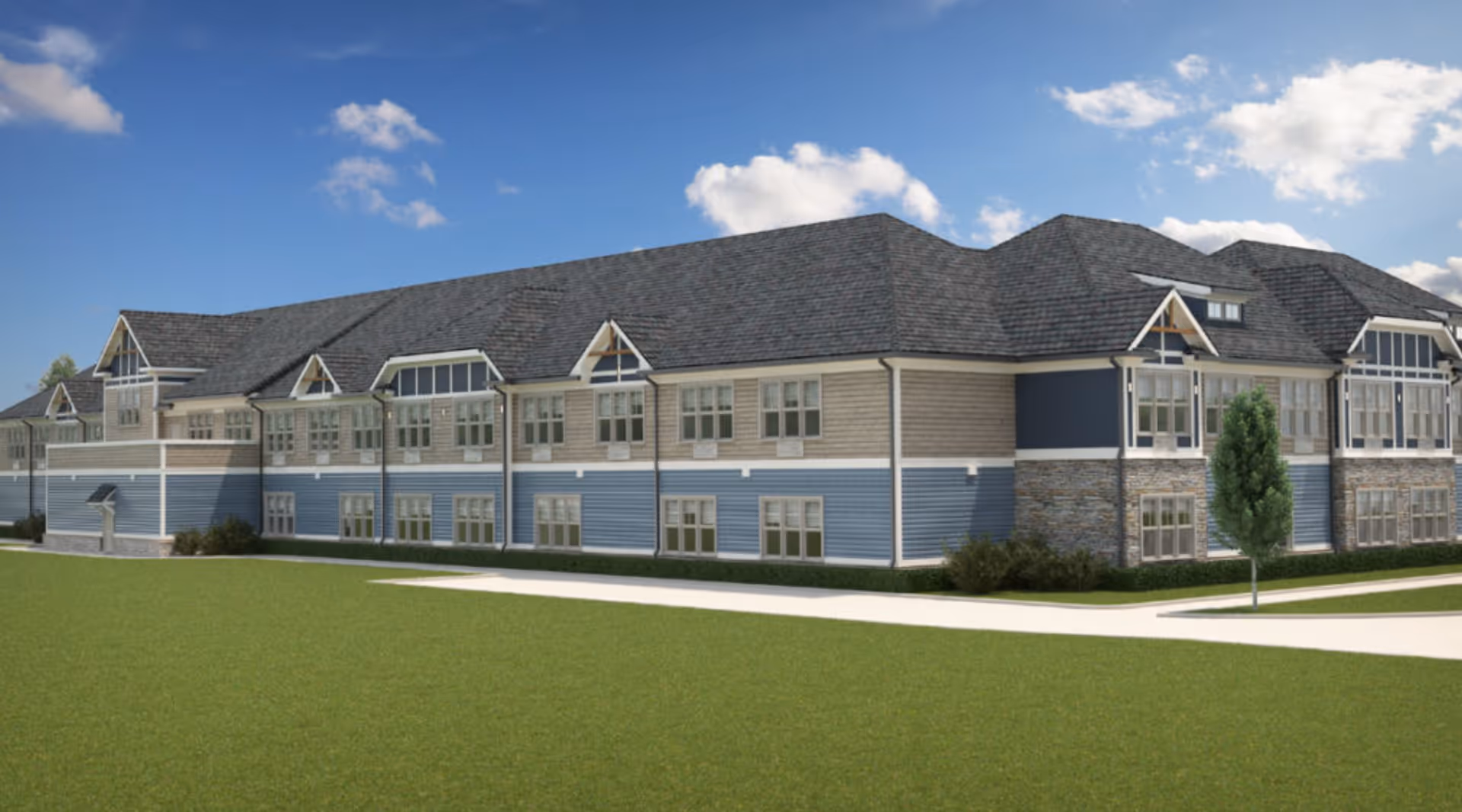 Exterior view of a large two-story senior living facility building with blue and beige siding, multiple windows, a dark shingled roof, and a small tree in front, under a partly cloudy blue sky.