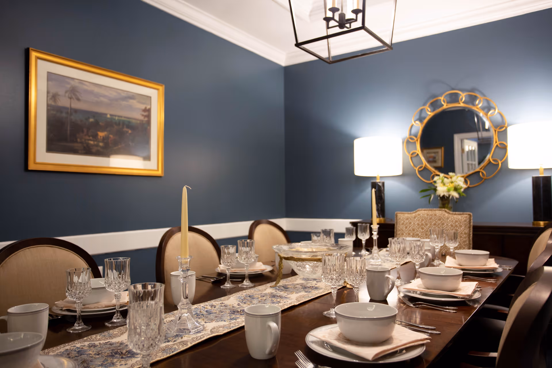 A formal dining room with a long wooden table set for a meal. The table has crystal glassware, white bowls, plates, mugs, and silverware arranged neatly on a decorative table runner. The room features blue walls with white trim, a framed painting, two table lamps with white shades, and a round decorative mirror on the wall.