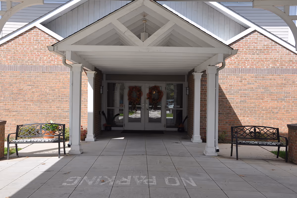 Covered entrance to a brick building with two benches on either side and double glass doors decorated with wreaths. The ground has a 'NO PARKING' sign painted on it.