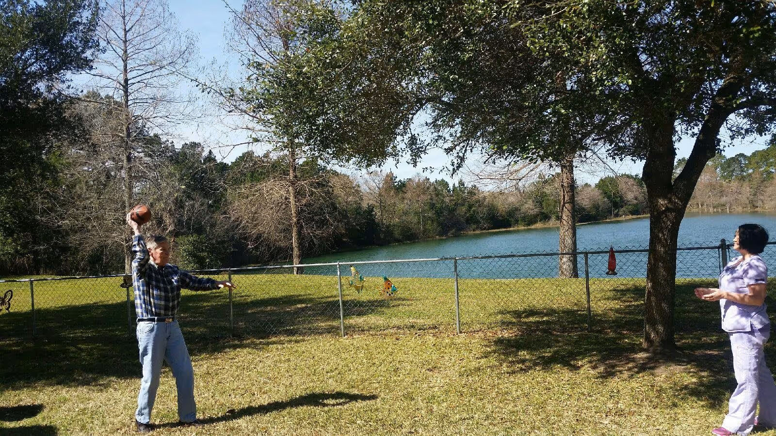 An elderly man and a woman standing outside near a chain-link fence with a lake and trees in the background. The man is holding a football, preparing to throw it, while the woman stands a few feet away watching him. The scene is set in a grassy area with some shade from nearby trees.