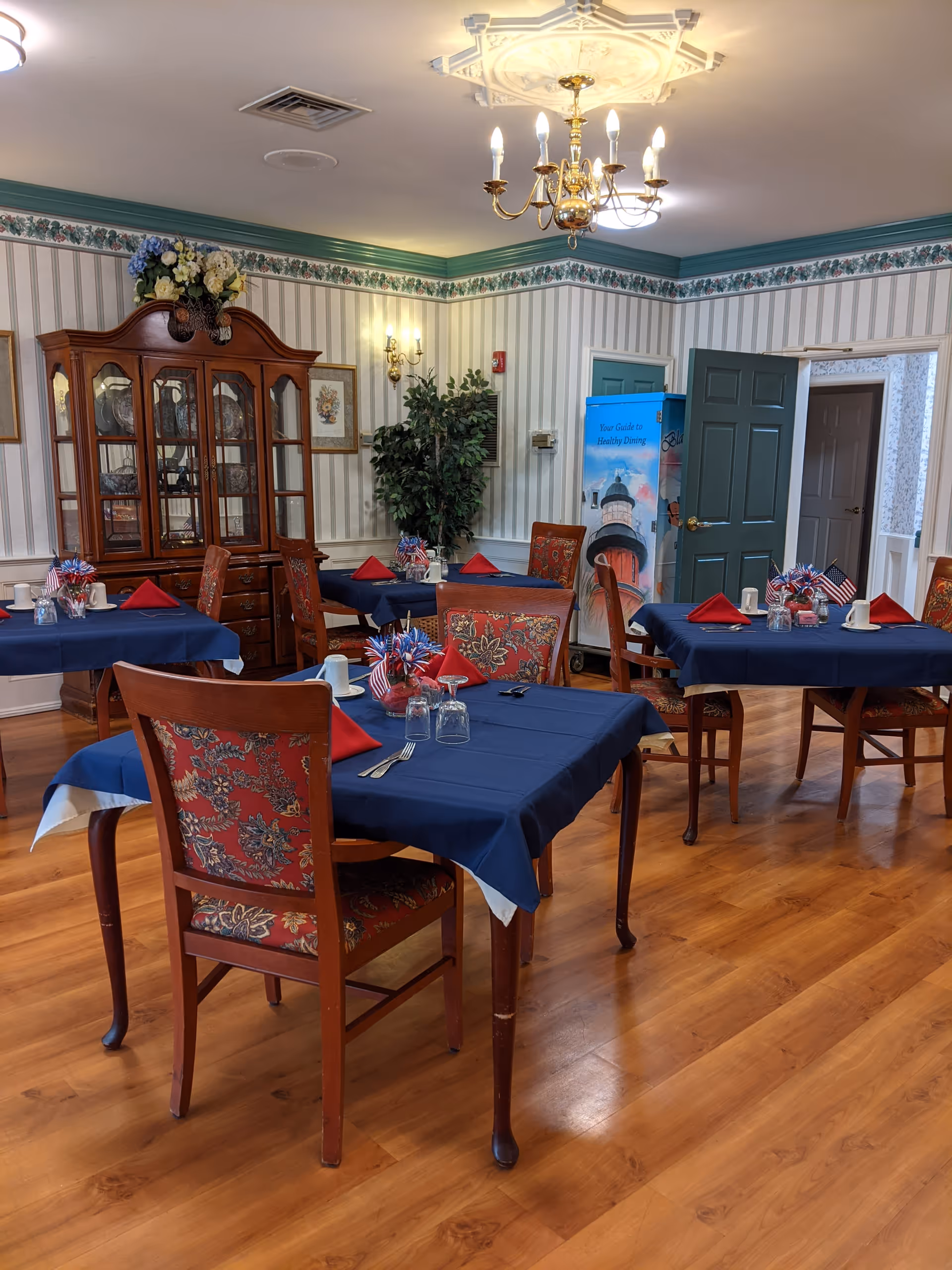 A dining room in an assisted living facility with wooden floors and several tables covered with blue tablecloths and red napkins. Each table has chairs with floral upholstery, small American flag decorations, and place settings including glasses and utensils. The room features striped wallpaper with a floral border near the ceiling, a wooden china cabinet with glass doors, a chandelier, wall sconces, and a potted plant in the corner. There is a blue door partially open and a poster with a lighthouse image in the background.