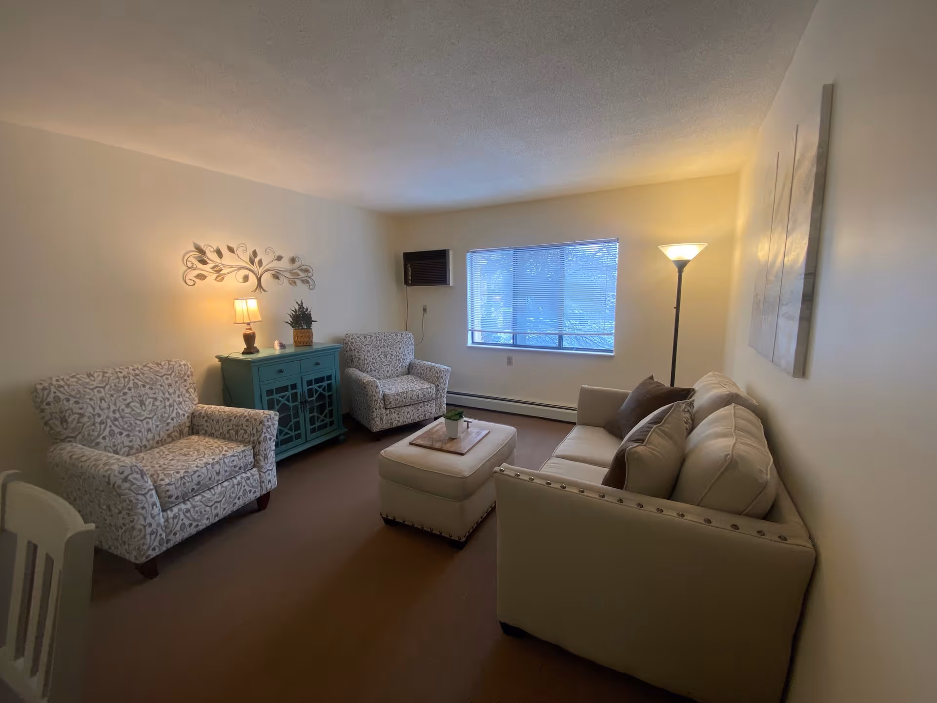 A cozy living room with a beige sofa adorned with brown pillows, two patterned armchairs, a teal cabinet with a lamp and plant on top, a small ottoman with a tray and plant, a floor lamp, and a window with blinds letting in natural light.