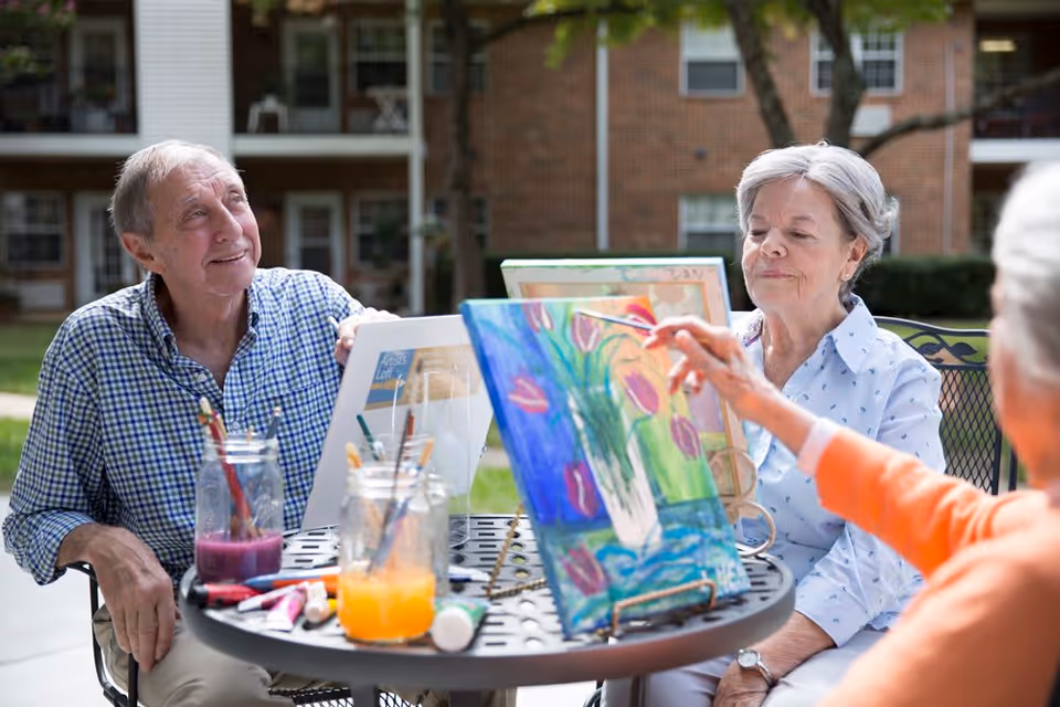 Three elderly people sitting outdoors around a round metal table, painting on canvases with brushes and colorful paints. They are engaged in an art activity with a brick building and greenery in the background.