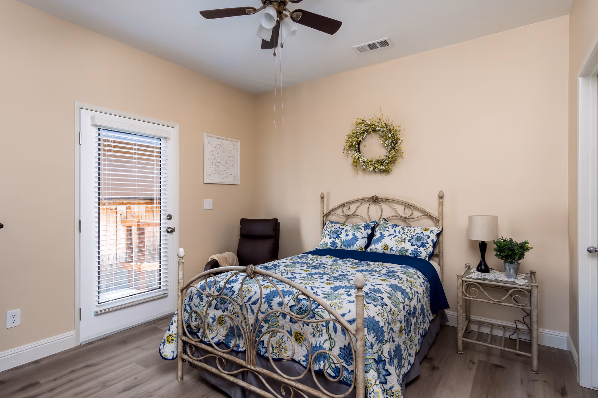 A cozy bedroom with a decorative metal bed frame, floral bedding in blue and white, a small side table with a lamp and a potted plant, a dark brown armchair in the corner, a ceiling fan with lights, and a door with blinds leading outside. The walls are painted beige and there is a decorative wreath hanging above the bed.
