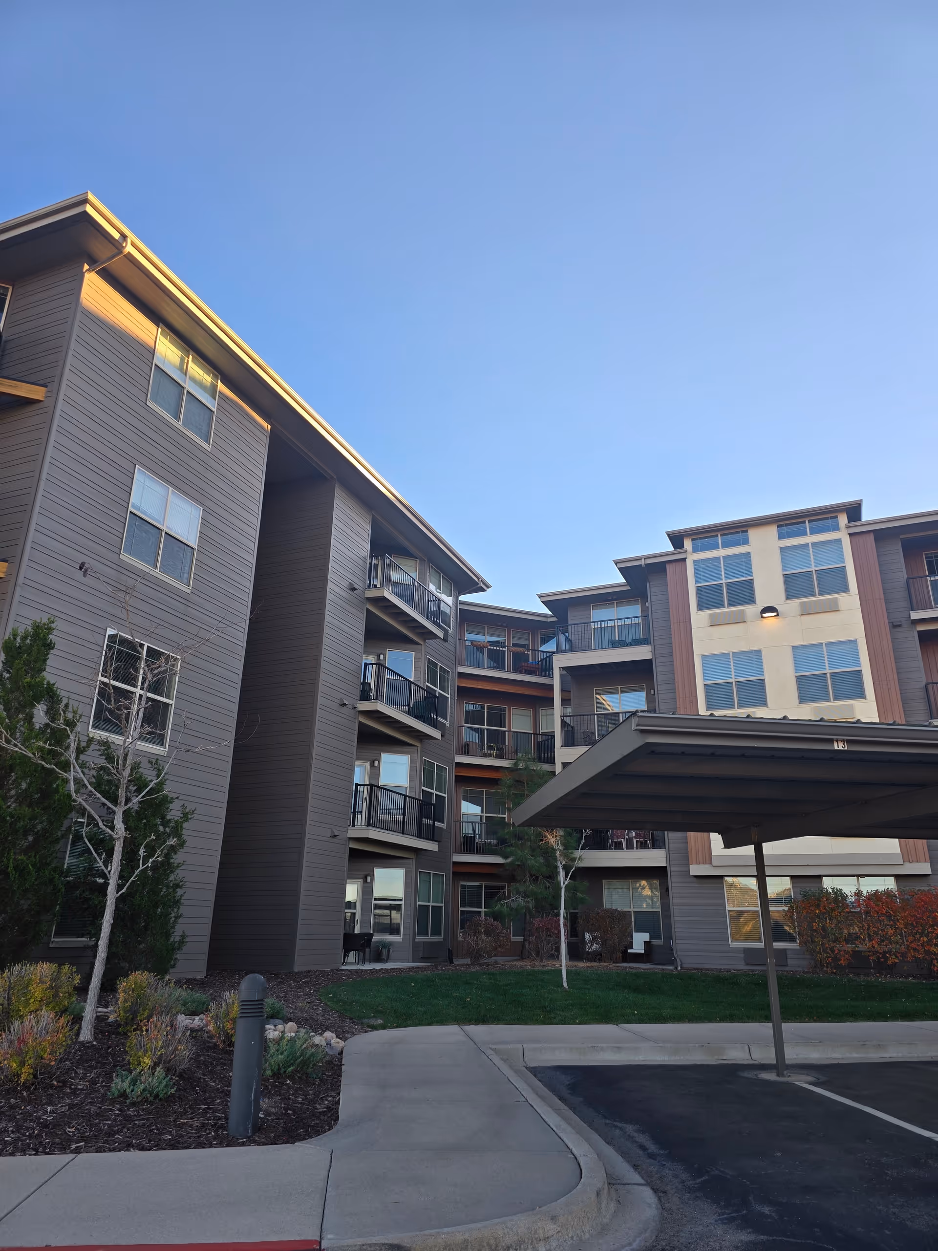 Exterior view of a multi-story residential building with balconies, surrounded by landscaped greenery and a covered parking area under a clear blue sky.