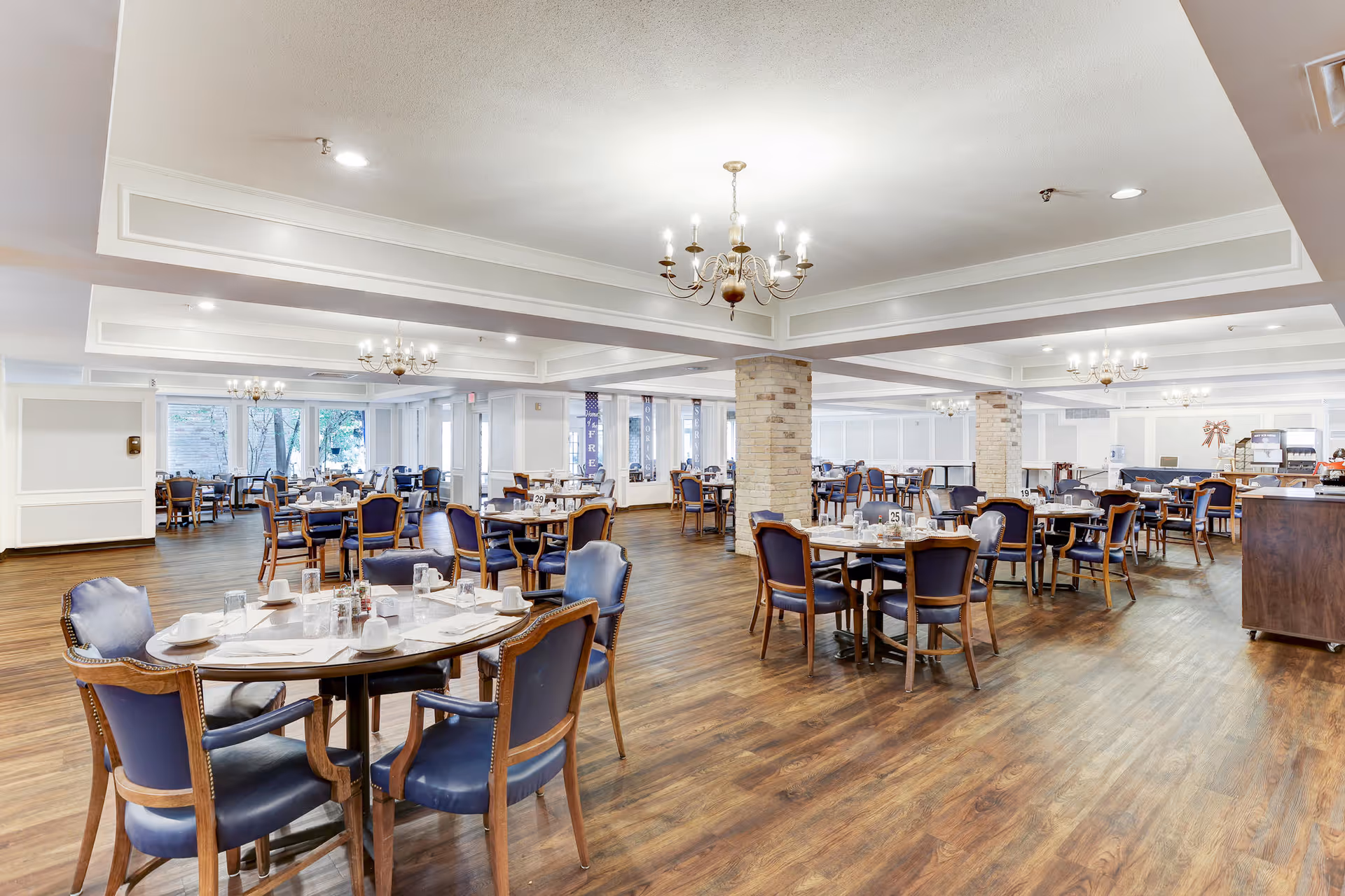 A spacious dining room in Treemont Senior Living with multiple round tables and blue cushioned chairs arranged neatly. The room features wooden flooring, white walls, brick columns, and several chandeliers hanging from the ceiling. The tables are set with white cups, glasses, and napkins, ready for dining.