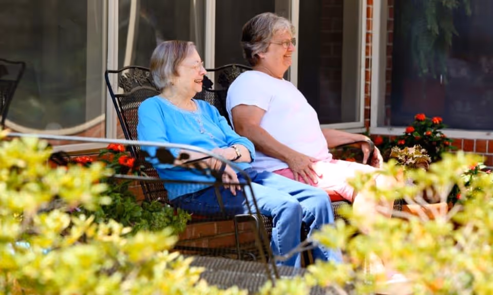 Two elderly women sitting and smiling on metal chairs outside a building, surrounded by plants and flowers.