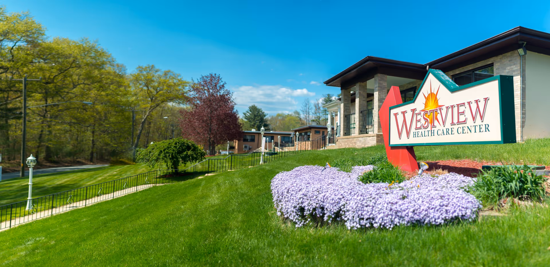 Entrance and front lawn of Westview Health Care Center featuring the facility sign, purple flowers, and the building facade.
