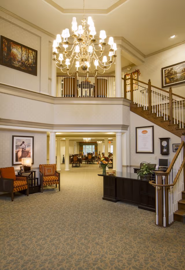 Interior view of a senior living facility lobby with a large chandelier hanging from the ceiling. There are two armchairs with orange cushions and a small table with a lamp on the left side. A dark wooden reception desk with a plant and flowers is on the right near a staircase with wooden handrails. The background shows a dining area with tables and chairs. The walls are decorated with framed paintings and a clock.
