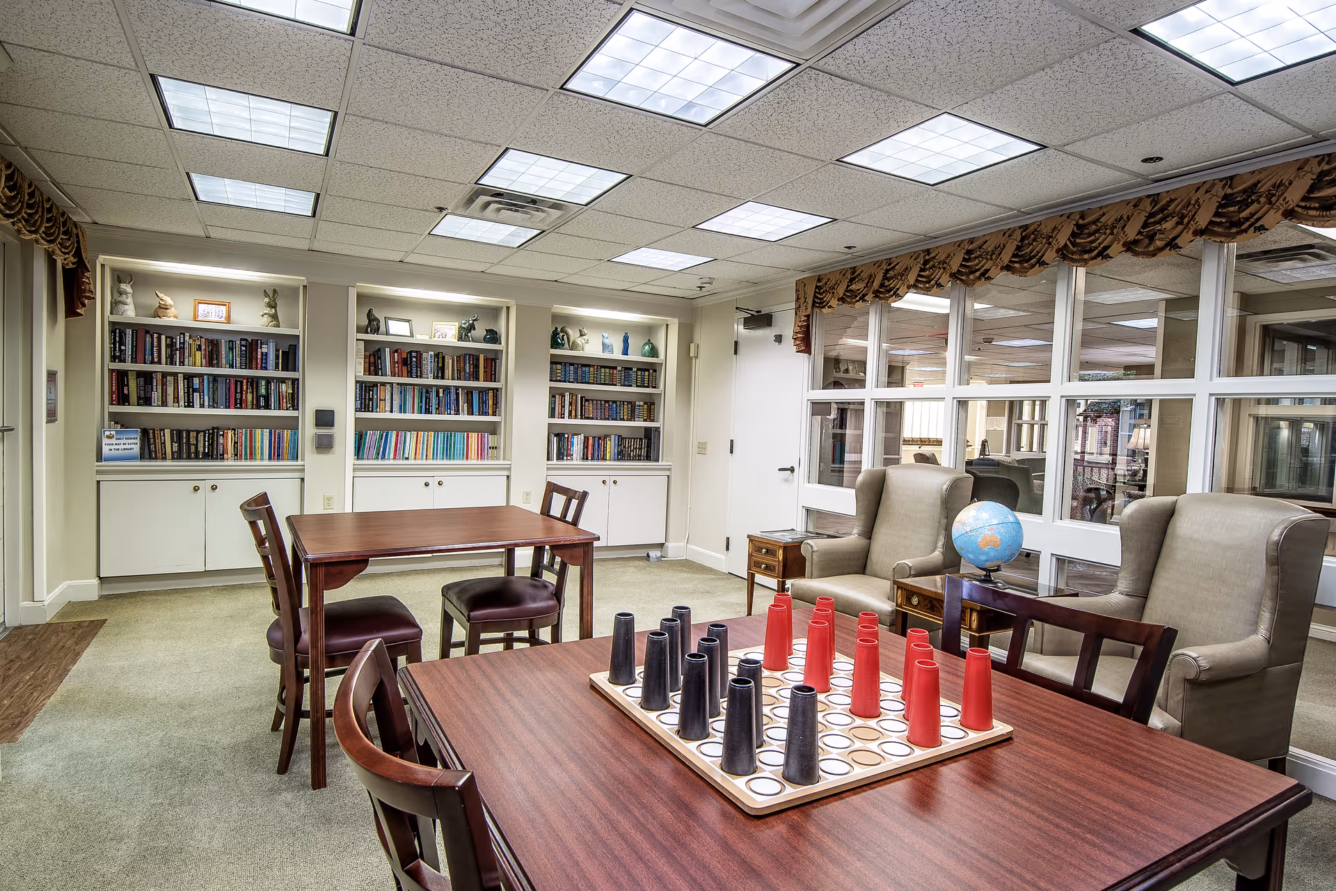 A well-lit communal library/lounge with tables and chairs, bookshelves, armchairs and a tabletop game.