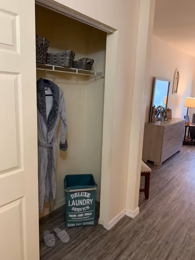 Open closet with a hanging gray robe, a green laundry bin labeled "Deluxe Laundry Service" and slippers beside a hallway leading to a dresser and lamp.