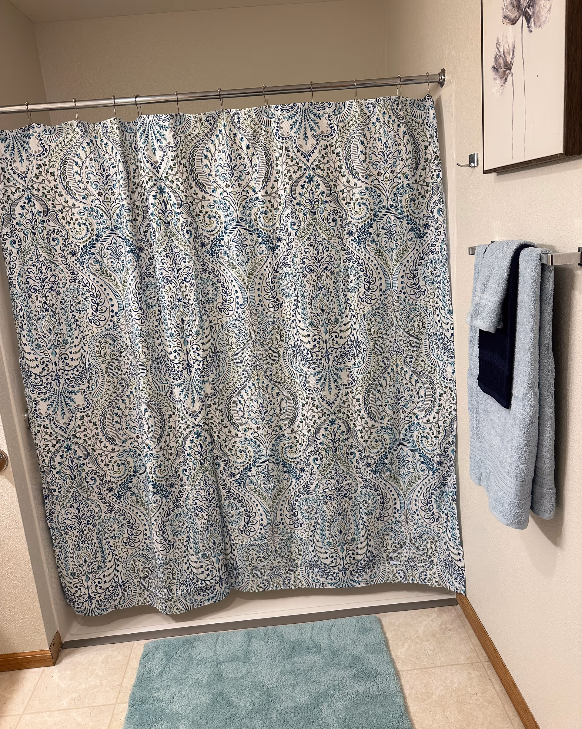 A bathroom with a patterned shower curtain featuring blue and green intricate designs. There are two towels hanging on a towel rack on the wall to the right, one light blue and one dark blue. A light blue bath mat is on the floor in front of the shower. A framed floral artwork is partially visible on the wall above the towel rack.