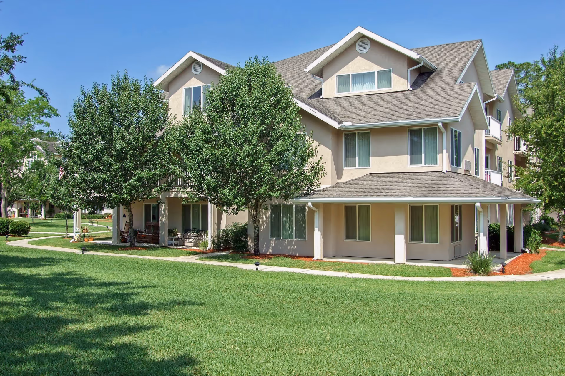 Exterior view of a multi-story beige senior living building with a covered porch, trees, and a manicured lawn.