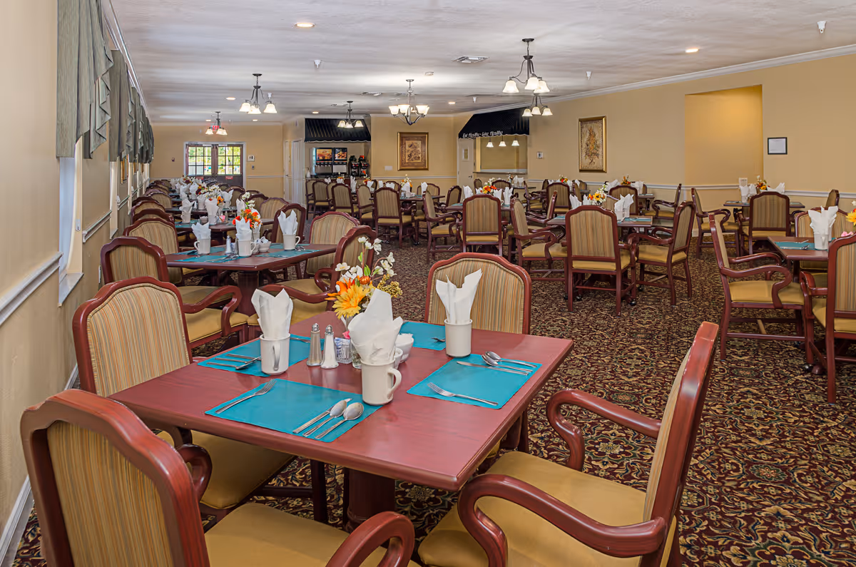 A spacious dining room with multiple wooden tables and cushioned chairs arranged neatly. Each table is set with turquoise placemats, white napkins folded in cups, silverware, salt and pepper shakers, and small floral centerpieces. The room has beige walls, patterned carpet, and several ceiling light fixtures providing warm lighting. Windows with green valances line one side of the room.