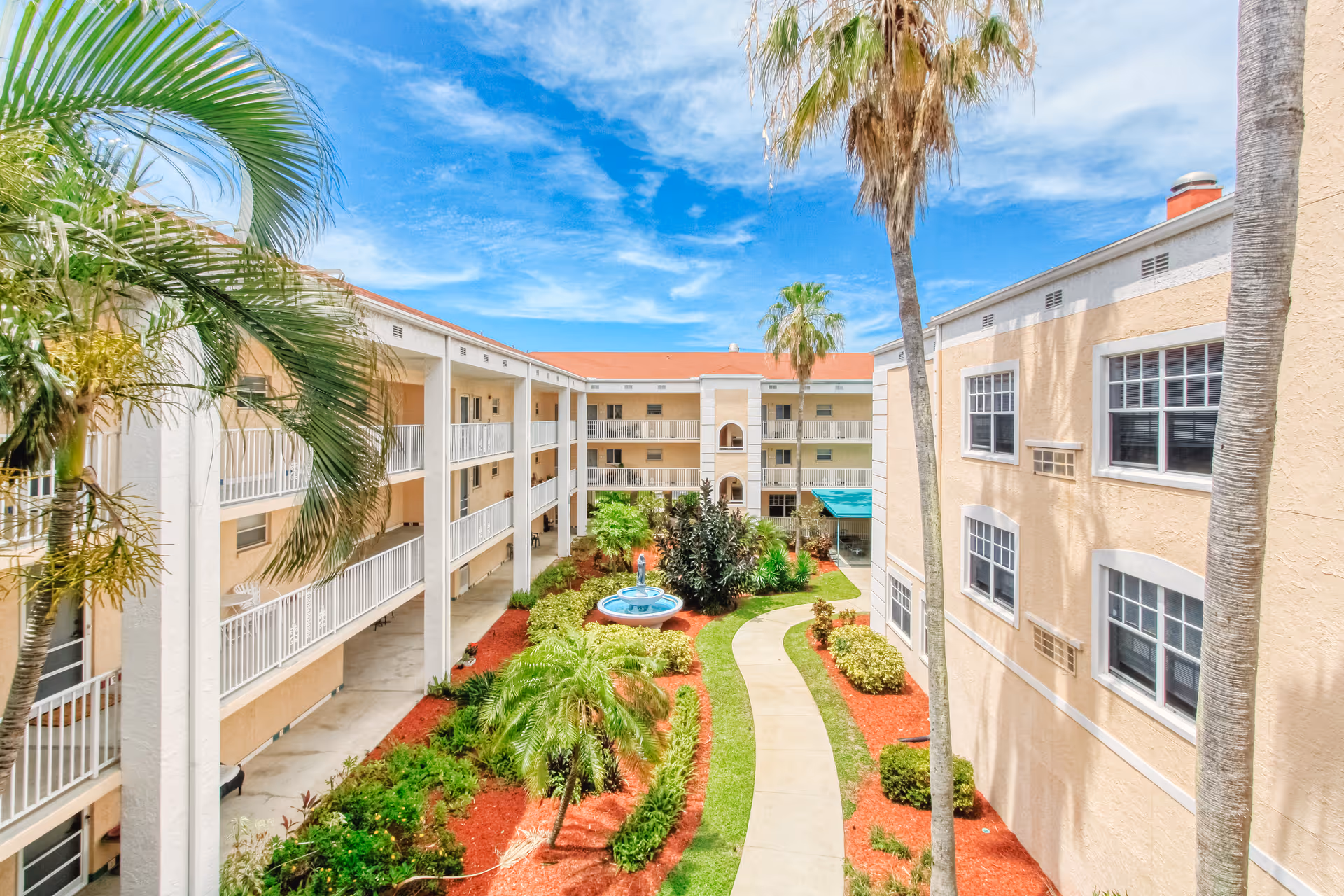 View of a sunny courtyard in a senior living facility with a paved walkway, green grass, palm trees, and a blue fountain in the center. The courtyard is surrounded by beige multi-story buildings with balconies and windows under a bright blue sky with some clouds.