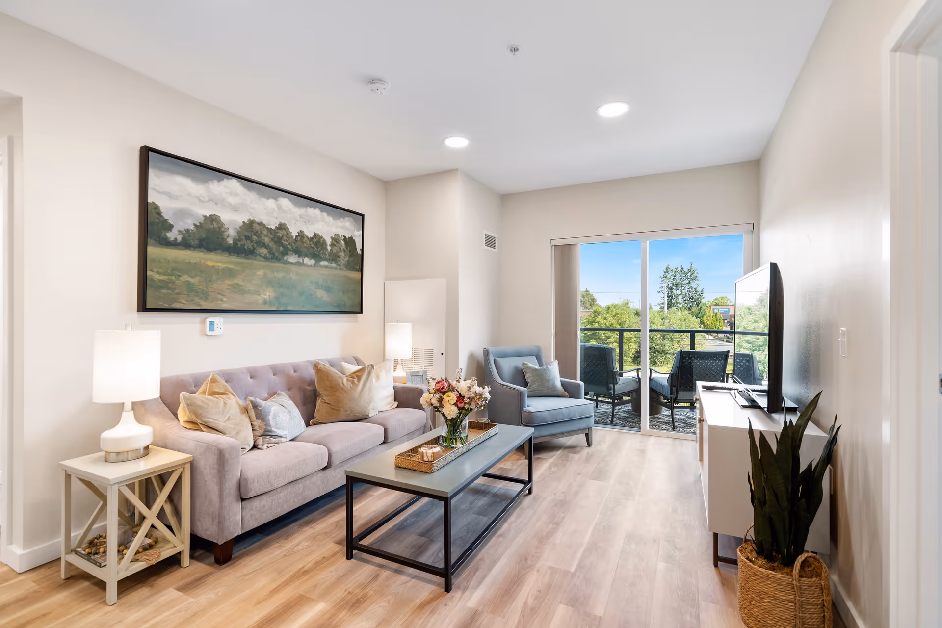 A bright and modern living room with a light gray sofa adorned with multiple pillows, a blue armchair, a rectangular coffee table with a flower arrangement, and a TV on a stand. The room has light wood flooring, a large landscape painting on the wall, two table lamps, and a sliding glass door leading to a balcony with outdoor seating and greenery visible outside.