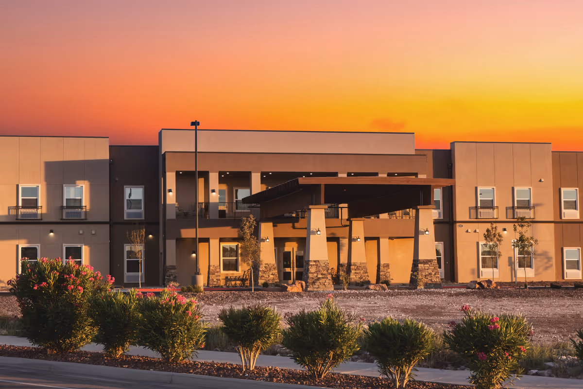 Exterior view of a modern two-story building at sunset with a covered entrance supported by stone pillars, surrounded by landscaped bushes and small trees.