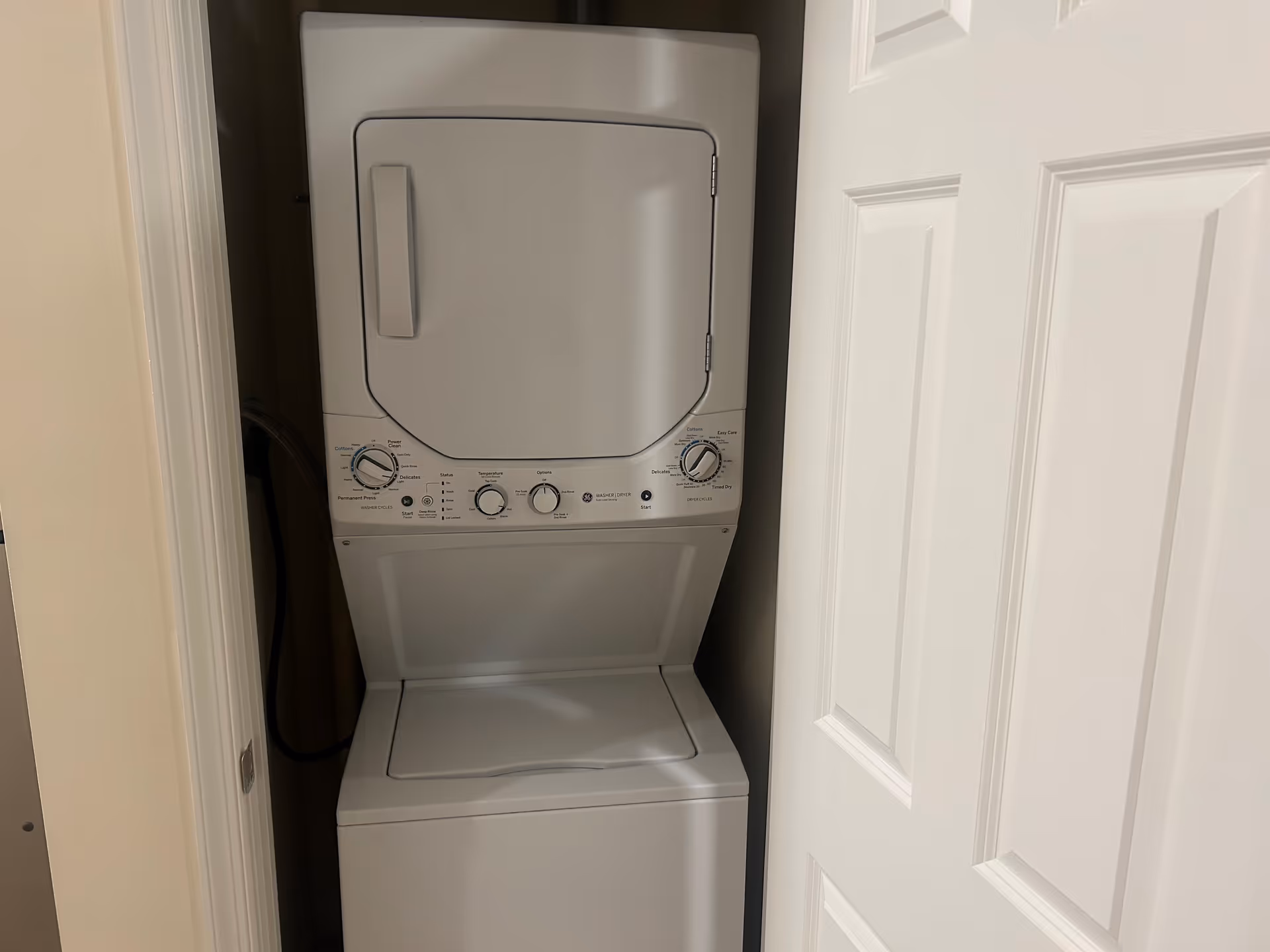 A stacked white washer and dryer unit inside a small laundry closet with a partially open white door.