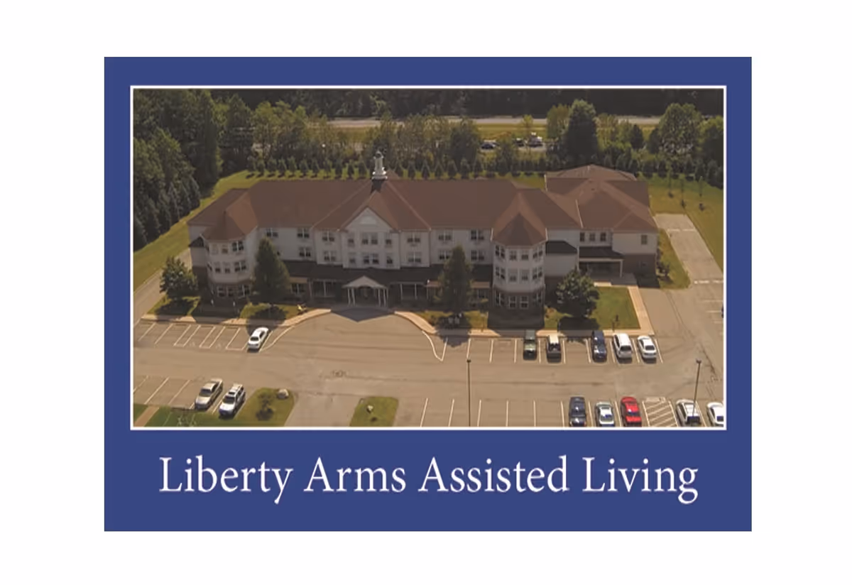 Aerial view of a large assisted living facility building with a brown roof, surrounded by parking lots with several cars and green trees in the background. The image is bordered by a blue frame with the text 'Liberty Arms Assisted Living' at the bottom.