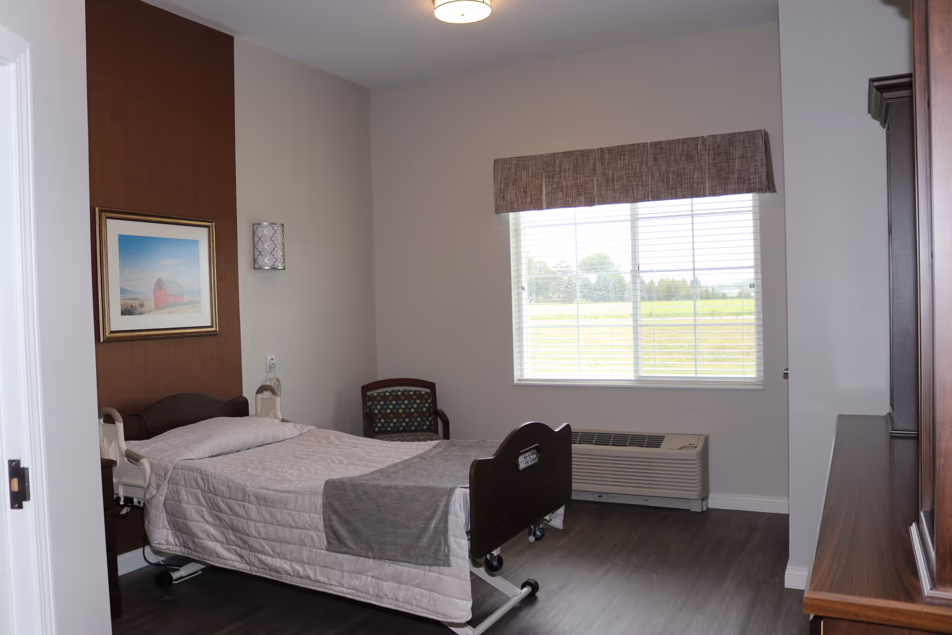 A senior living bedroom with an adjustable hospital-style bed, a chair, window with blinds, and wood furniture.