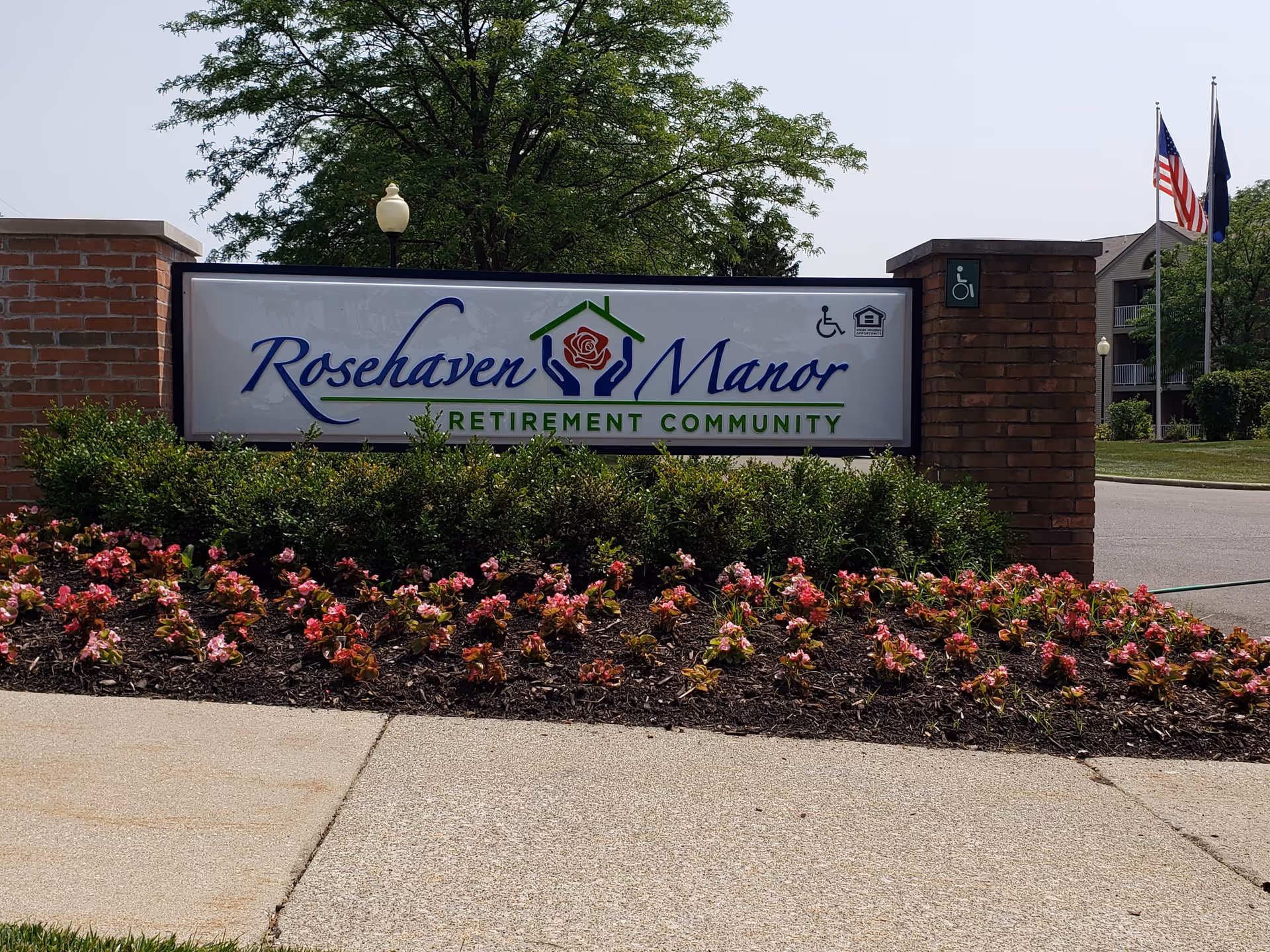 Entrance sign for Rosehaven Manor Retirement Community surrounded by flower beds and greenery, with American and state flags in the background.