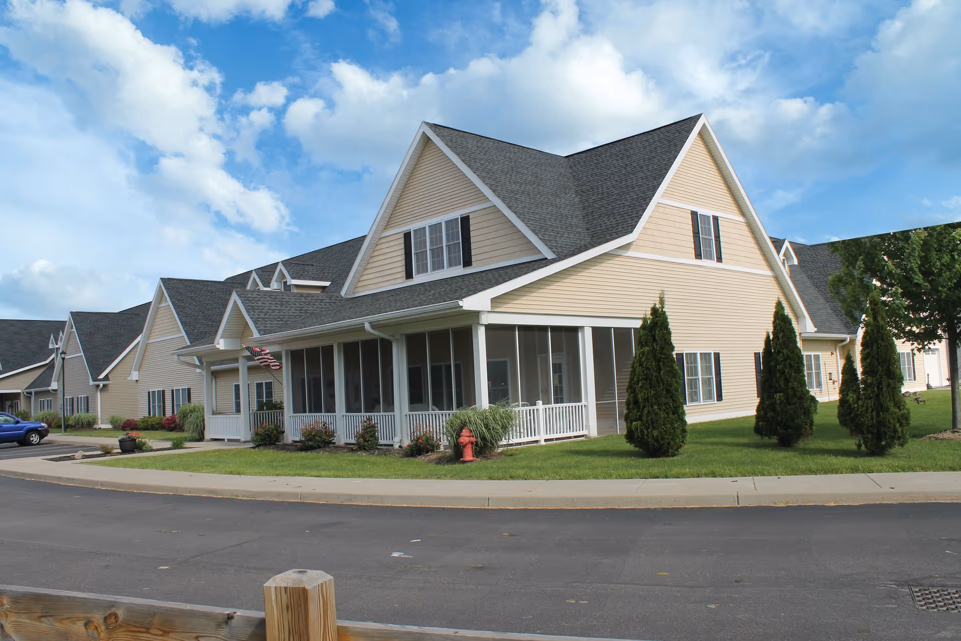 Exterior view of a beige senior living facility building with multiple peaked roofs, white trim, and a screened porch. The building is surrounded by a well-maintained lawn, small trees, shrubs, and a paved road in front. The sky is partly cloudy.