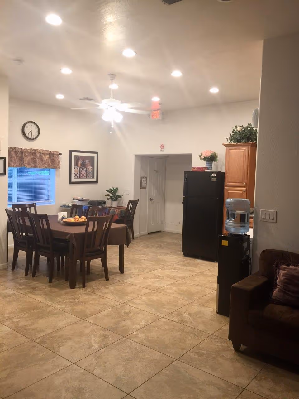 Interior view of a dining area in an assisted living facility featuring a rectangular dining table with six chairs, a ceiling fan with lights, a wall clock, framed pictures, a window with a valance, a black refrigerator, wooden cabinets, a water dispenser, and a brown couch with a purple pillow.