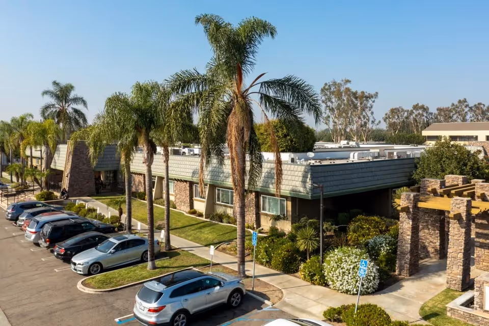 Exterior view of a single-story senior living facility building with a row of parked cars in front, palm trees lining the sidewalk, and a stone pergola structure on the right side under a clear blue sky.