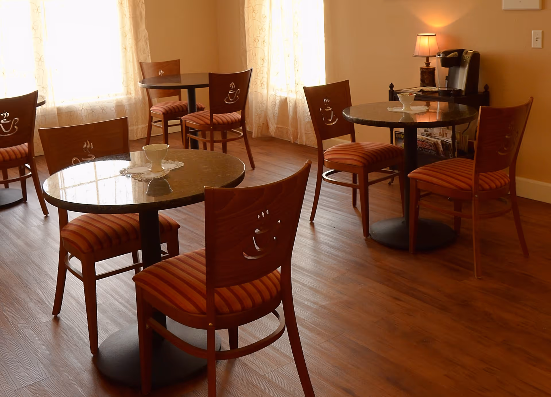 A cozy dining area with round tables and wooden chairs featuring striped cushions and coffee cup designs on the backrests. The room has warm lighting, sheer curtains on the windows, and a coffee station with a lamp in the corner.