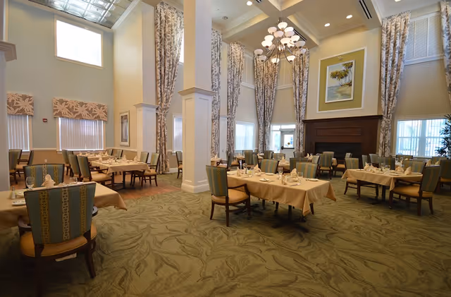 A spacious dining room with high ceilings, large windows with patterned curtains, multiple tables covered with beige tablecloths, and chairs arranged neatly around the tables. The room features a large chandelier, a fireplace with a framed painting above it, and a carpeted floor with a leafy pattern.
