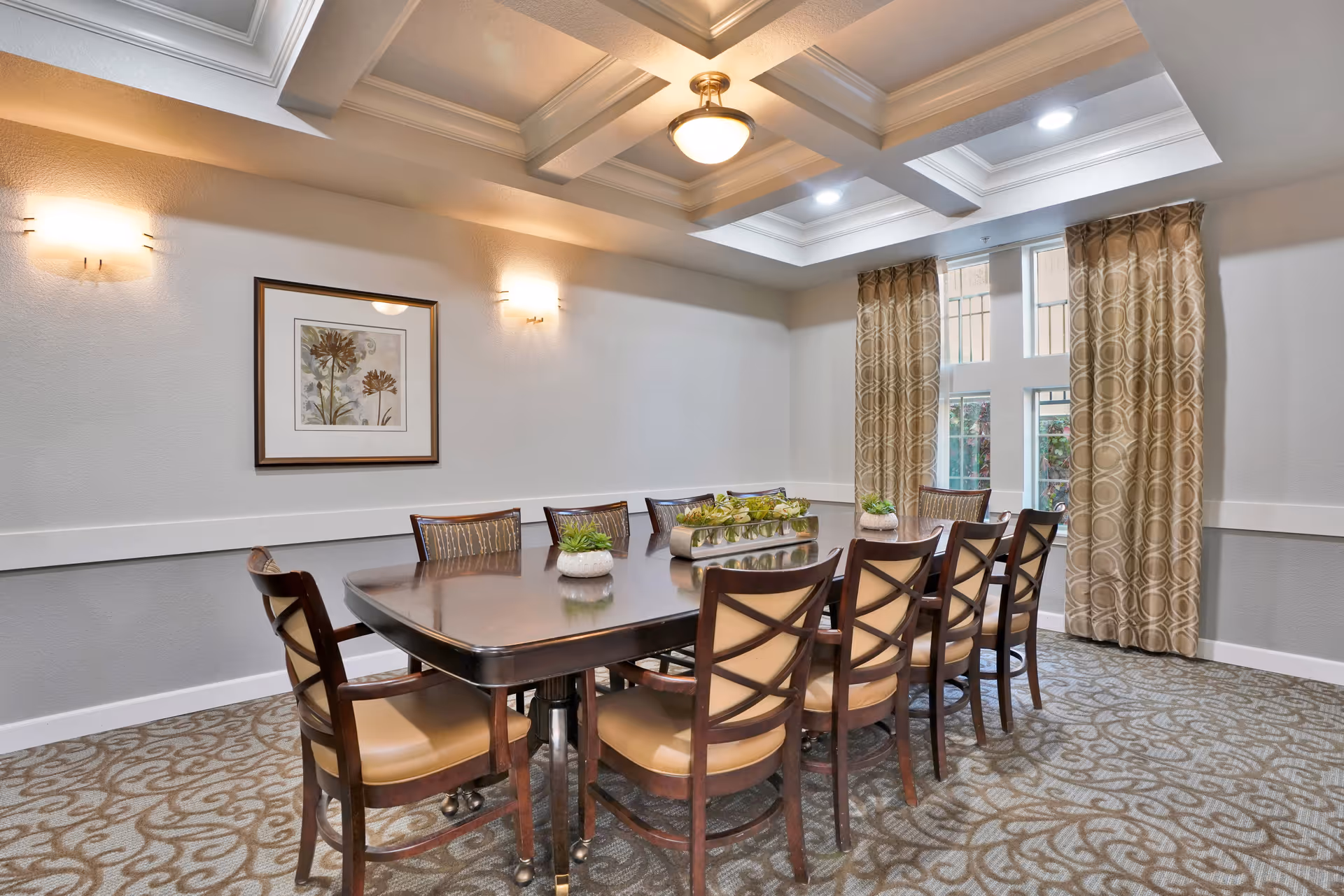A well-lit dining room with a large rectangular wooden table surrounded by ten cushioned chairs. The room features patterned carpet, beige walls with white trim, a framed floral artwork on the wall, and large windows with patterned curtains allowing natural light to enter. The ceiling has recessed lighting and a central light fixture.