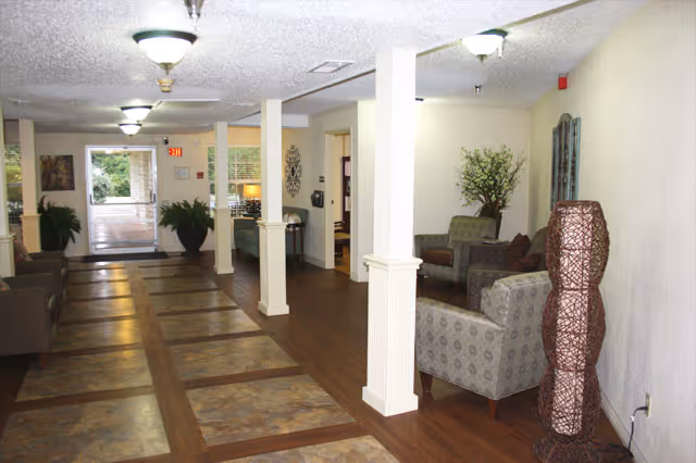 Interior view of a nursing and rehabilitation center hallway with seating areas. The hallway has a tiled floor with wooden borders, white pillars, and ceiling lights. There are several armchairs and a decorative lamp on the right side, along with plants and wall decorations. At the far end, there is a glass door leading outside.