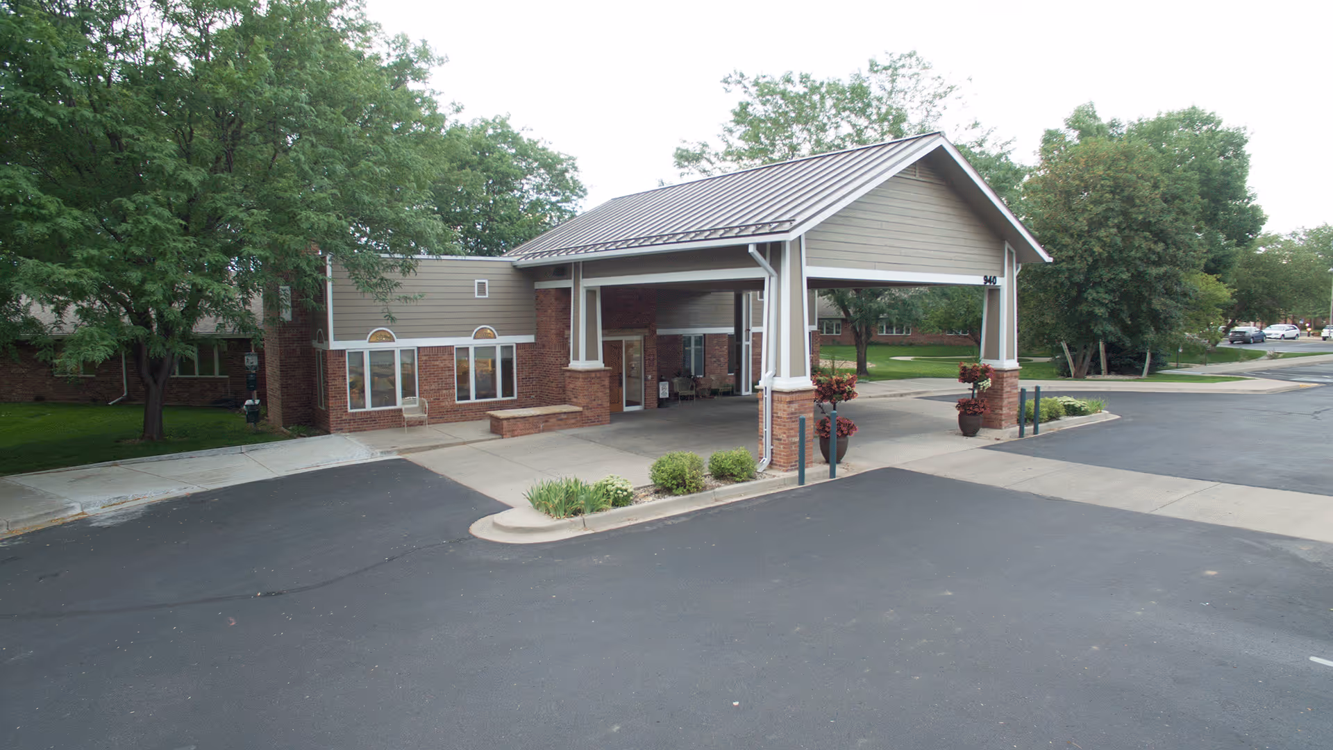 Front exterior view of Columbine West Health & Rehab Facility showing a covered entrance with a peaked roof supported by columns, brick and siding walls, surrounding greenery, and a paved driveway and parking area.