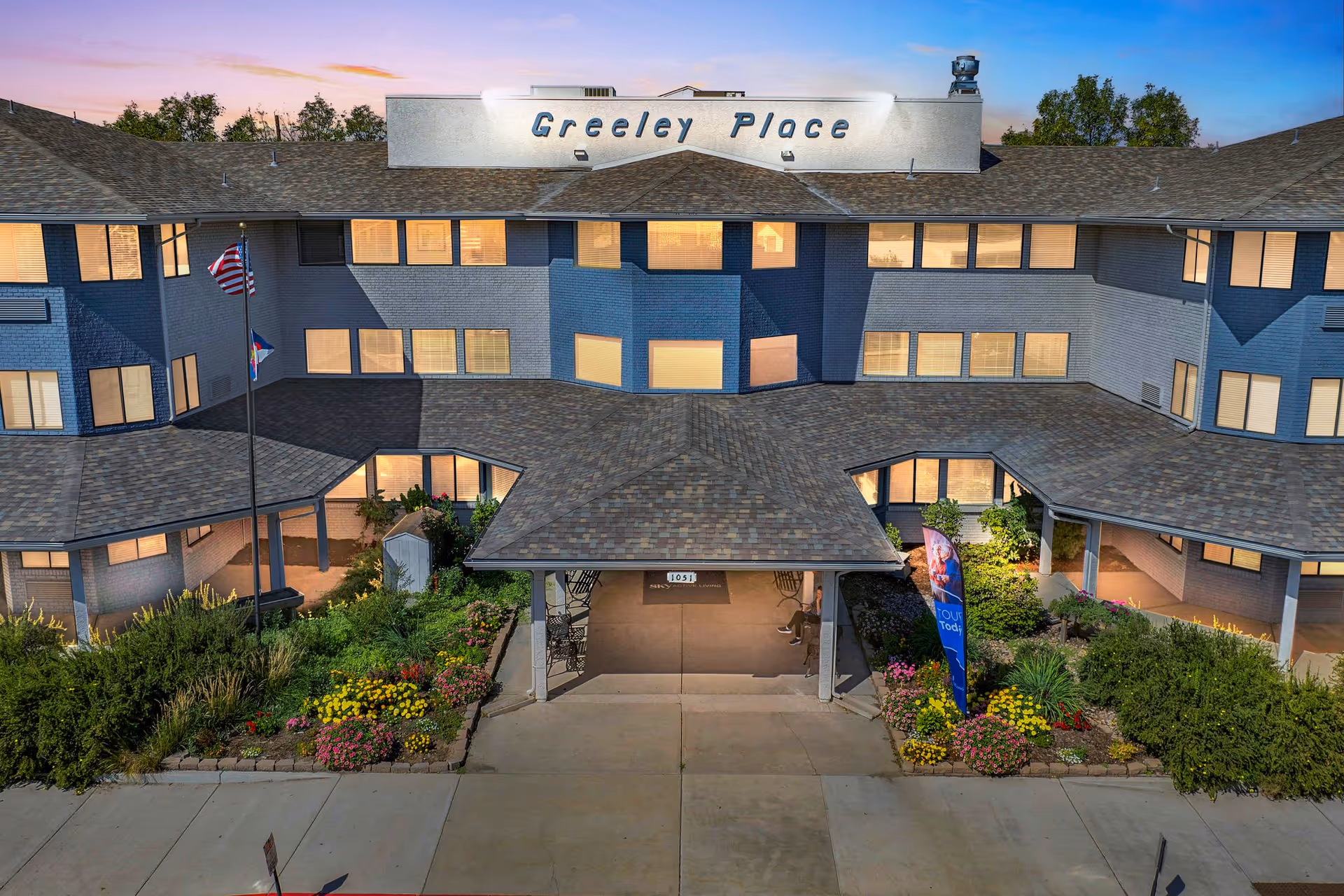 Exterior view of Greeley Place senior living facility at dusk, showing a three-story building with many lit windows, a covered entrance, landscaped flower beds, and two flagpoles with flags.
