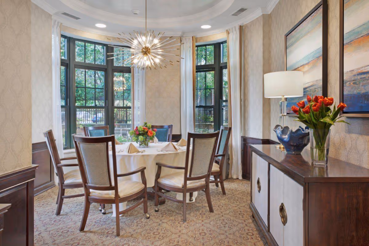 A bright dining room with a round table covered with a white tablecloth and set with napkins and silverware. Six wooden chairs with cushioned seats surround the table. Large windows with white curtains allow natural light to fill the room. A modern chandelier hangs above the table. A sideboard with a lamp, a blue decorative bowl, and a vase of red tulips is positioned against the wall, which is adorned with two abstract paintings.