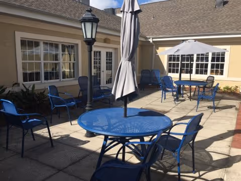 Sunlit courtyard patio with blue metal tables and chairs, umbrellas, and surrounding building windows.