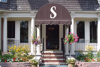 Entrance of a building with a brown awning displaying a large white letter 'S'. The entrance is flanked by white columns and windows, with colorful flower arrangements and greenery in front.