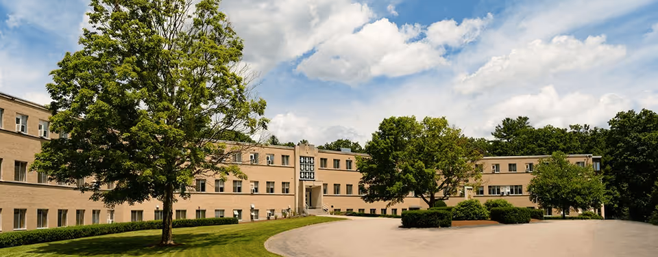 Exterior view of a large, three-story beige brick building with multiple windows, surrounded by green trees and bushes under a partly cloudy sky. A curved driveway leads to the entrance of the building.