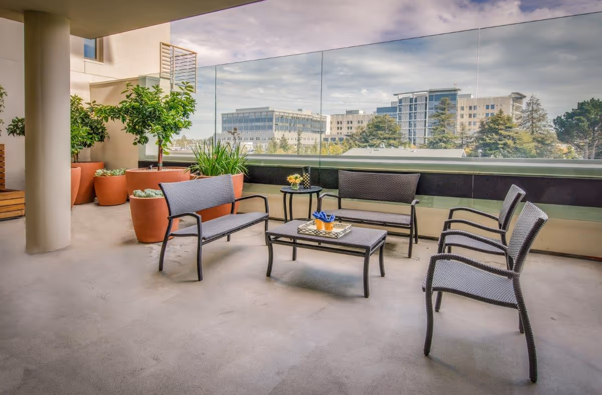 Outdoor patio area with modern wicker seating including two benches, three chairs, and a coffee table with decorative items. Large potted plants line the side, and a glass railing offers a view of nearby buildings and trees under a partly cloudy sky.