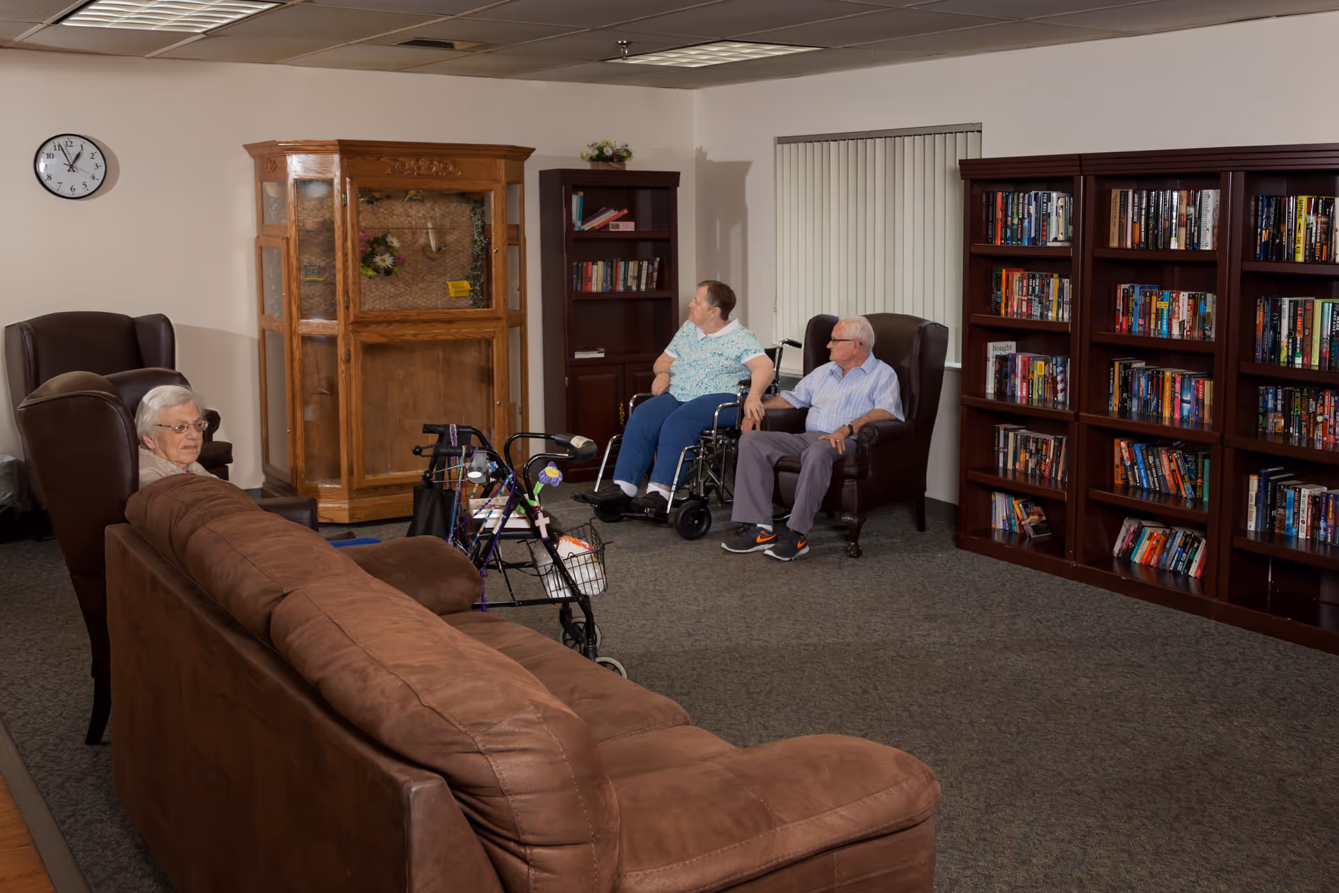 A cozy common room in a senior living facility with three elderly people seated. One woman is sitting on a brown leather chair near a brown couch, while another woman in a wheelchair and a man in a chair are near a wooden cabinet and bookshelves filled with books. The room has carpeted floors, a clock on the wall, and vertical blinds on the window.