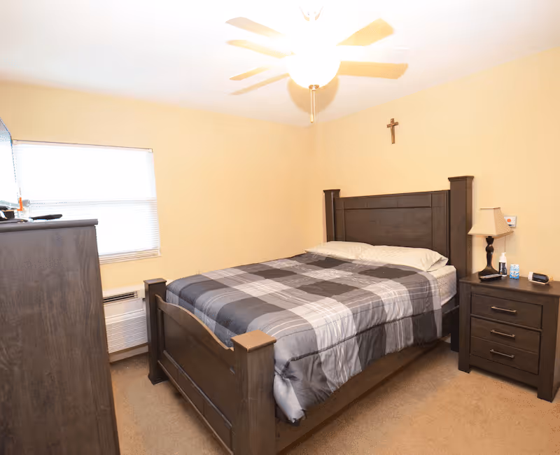 A bedroom with a wooden bed frame and a plaid comforter in shades of gray. There is a wooden nightstand with a lamp, a tissue box, and other small items. A wooden dresser is visible on the left side near a window with blinds. A ceiling fan with a light is on the ceiling, and a small cross hangs on the wall above the bed.