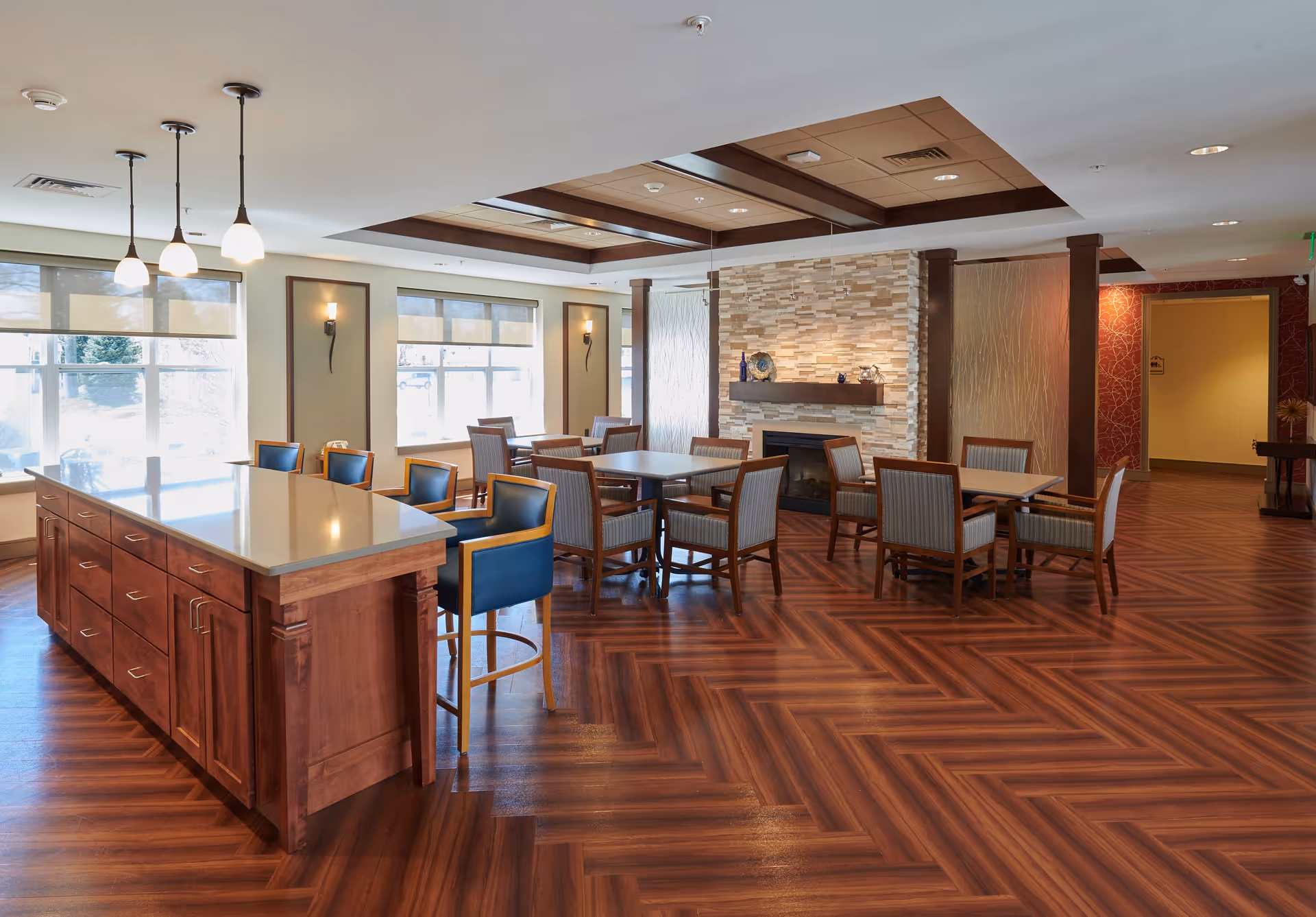 A spacious, well-lit dining area with wooden flooring and multiple tables and chairs. There is a large kitchen island with blue cushioned bar stools on one side. The room features large windows with blinds, a stone fireplace with decorative items on the mantel, and a ceiling with wooden beams and recessed lighting.