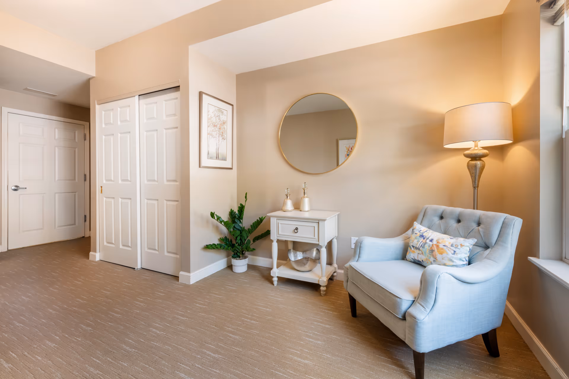 Neutral-toned sitting area with a light blue upholstered armchair, side table beneath a round mirror, floor lamp and potted plant.