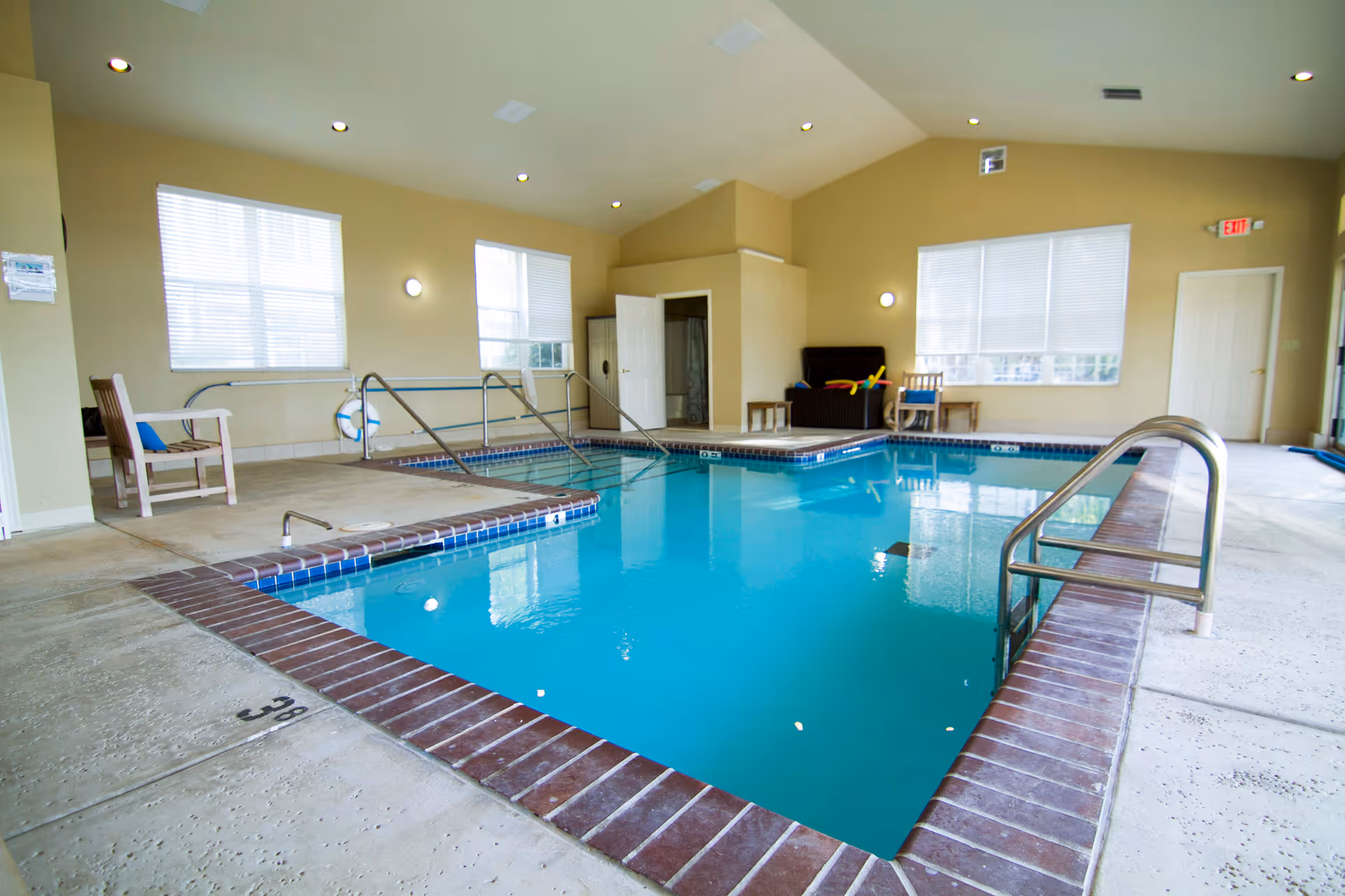 Indoor community swimming pool with metal handrails, seating, and large windows in a bright room.