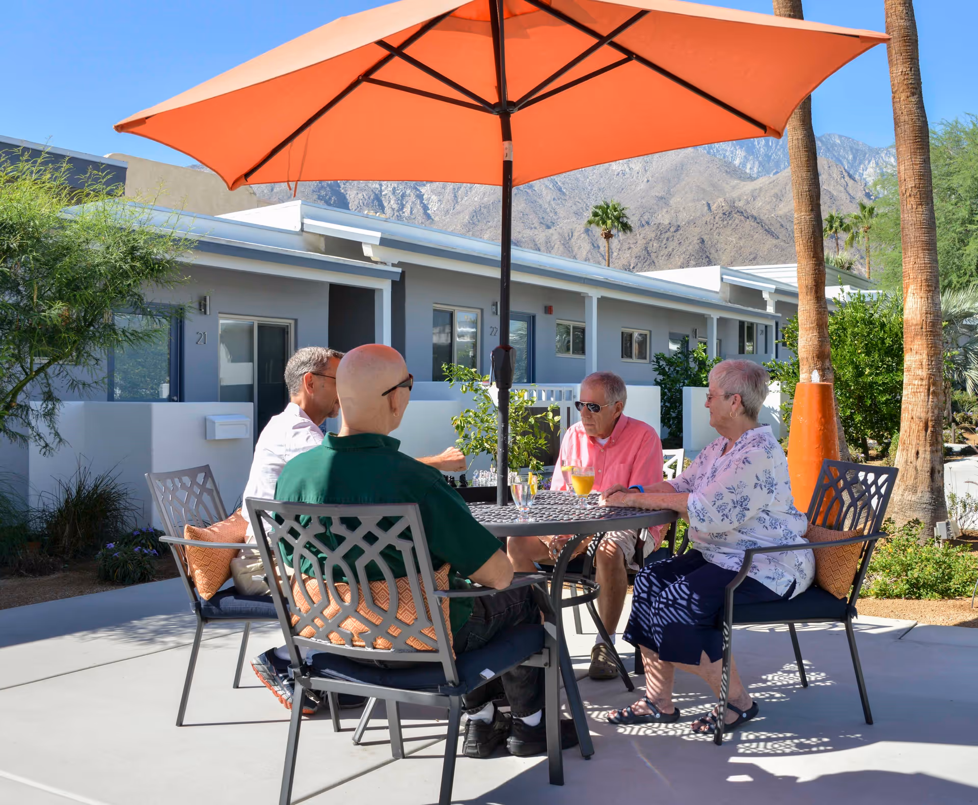 Four elderly people sitting around a metal outdoor table with an orange umbrella, enjoying drinks and conversation in a sunny courtyard with palm trees and mountains in the background.