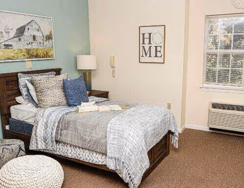 A furnished bedroom with a single bed, pillows and throw, bedside lamp and wall art, plus a window and wall-mounted air conditioner.