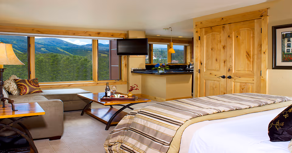 A cozy and well-lit living space featuring a bed with striped bedding in the foreground, a wooden coffee table with a bottle of wine and glasses, a beige sectional sofa with decorative pillows, a flat-screen TV mounted on the wall, large windows showing a scenic view of green hills and blue sky, wooden double doors, and a small kitchen area with a counter and pendant light.