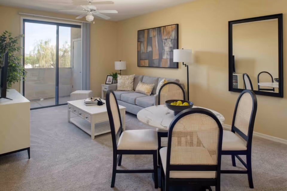 A cozy living room and dining area with a gray sofa, a white coffee table, a round dining table with four chairs, a large wall mirror, and a ceiling fan. There is a sliding glass door leading to a balcony with outdoor views and a potted plant near the TV stand.