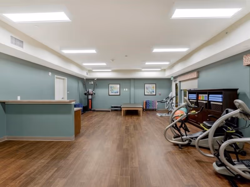 A spacious exercise room with wooden flooring and greenish walls. The room contains various fitness equipment including stationary bikes, an elliptical machine, a treadmill, and a rack with colorful exercise mats and weights. There is a counter on the left side and two framed pictures on the far wall. The ceiling has multiple rectangular fluorescent lights.
