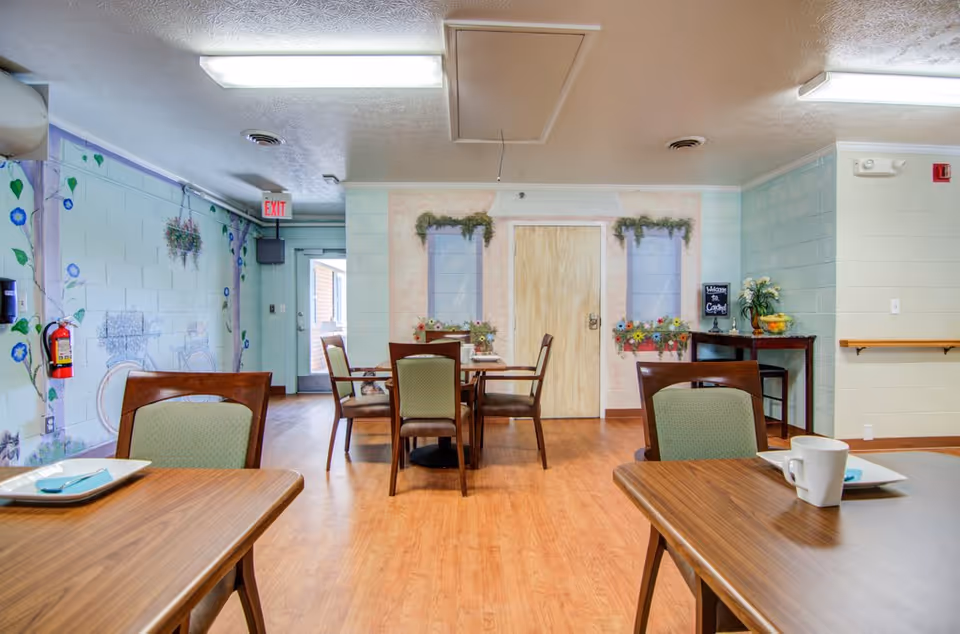 Interior view of a senior living facility dining area with wooden tables and chairs. The walls are decorated with murals of flowers and windows with flower boxes. There is a small table with a welcome sign, a bowl of fruit, and a vase of flowers. The floor is wooden, and fluorescent lights are on the ceiling.